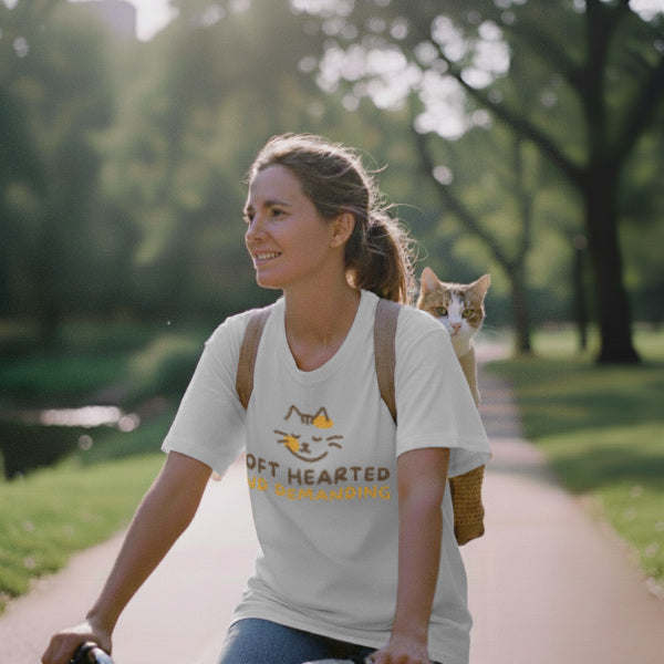 A woman in her 30s bikes during the day, wearing a white cotton t-shirt. Her cat rides along in a backpack, with its head visible peeking out from behind her.