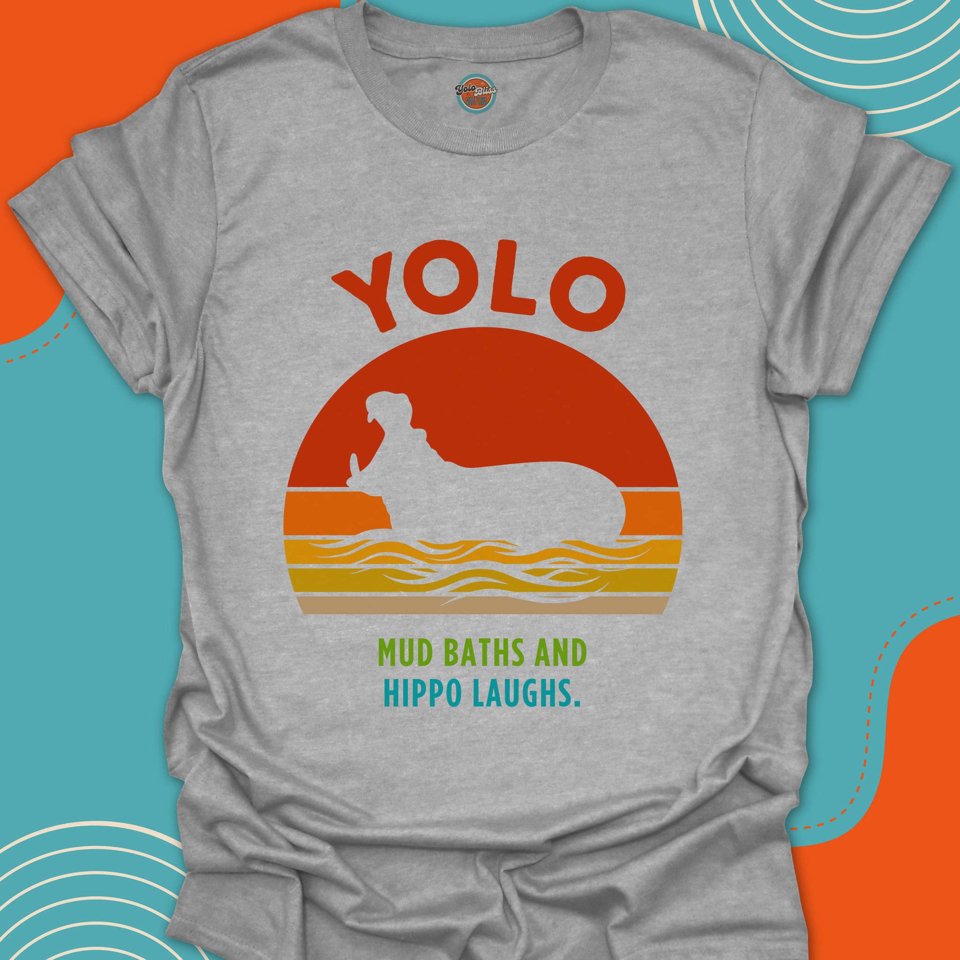 Light gray t-shirt with 'YOLO Mud Baths and Hippo Laughs' text and a hippopotamus with open mouth in a river design on a red orange and yellow sunset background