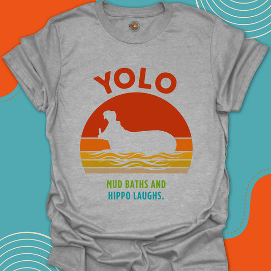 Light gray t-shirt with 'YOLO Mud Baths and Hippo Laughs' text and a hippopotamus with open mouth in a river design on a red orange and yellow sunset background